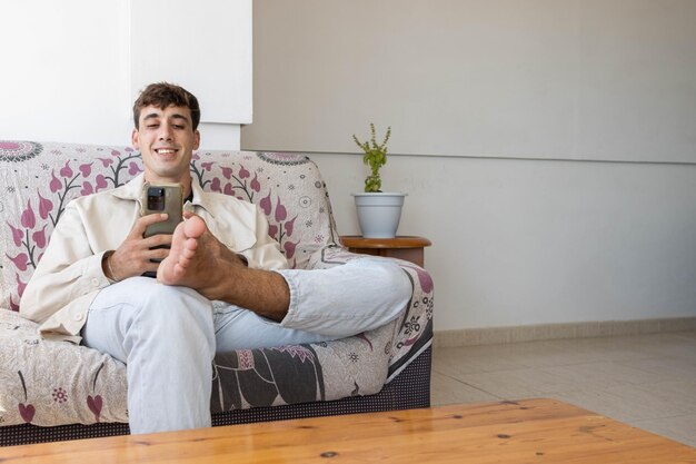 Young boy sitting on the sofa at home barefoot while looking at his mobile phone