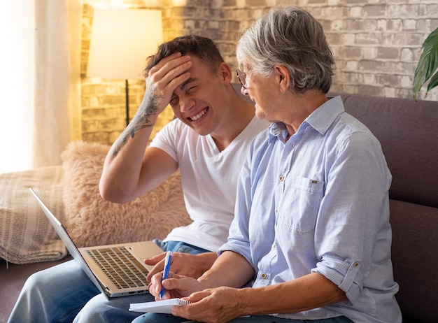 Young boy sitting on sofa at home helping senior grandmother\
surf with laptop on the web while she takes notes younger\
generations care for older relatives are teaching computers to\
use