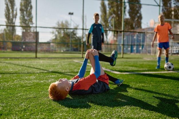 試合中にフィールドに横たわっている負傷した膝を持つ少年サッカー選手。ピッチで悪い一日を過ごしている苦痛で地面に足の痛みで子供のフットボール選手を傷つける