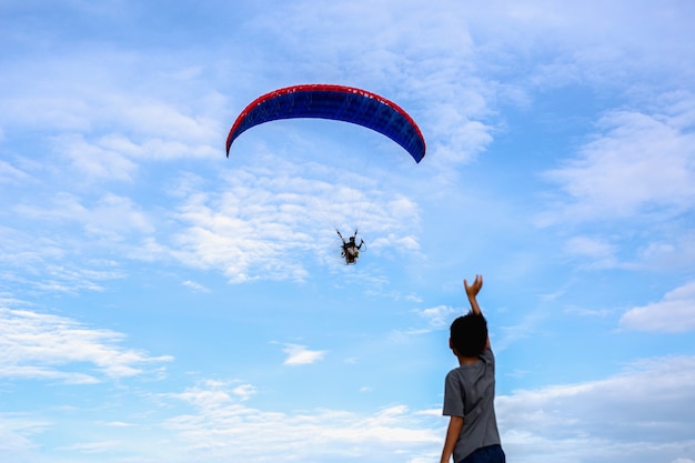 Young boy standing on the rock hands raised up looking at motor paraglider flying 