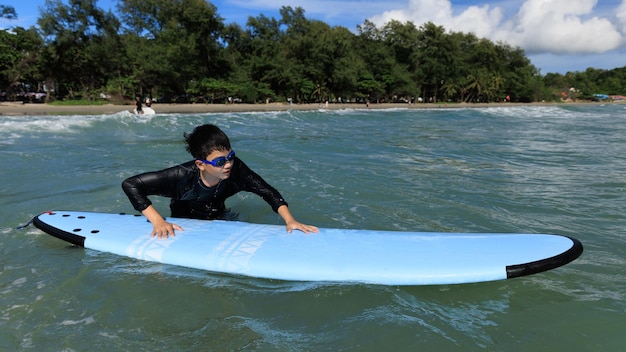 Young boy student in surfing is holding onto softboard and trying to bring it back into sea to practice while playing against waves and splashing water