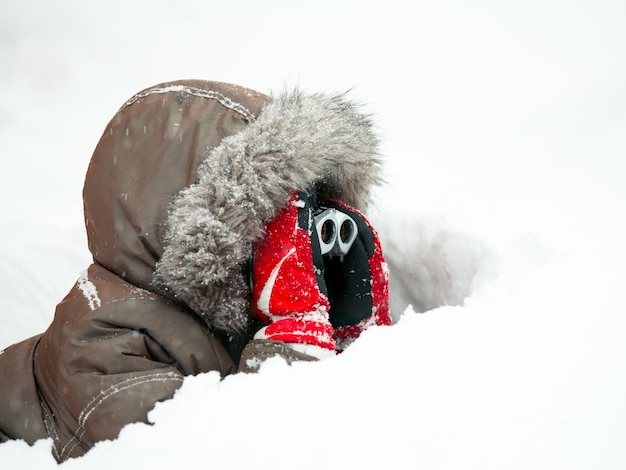 Молодой мальчик в зимней куртке с пушистым капюшоном и красными перчатками смотрит в бинокль