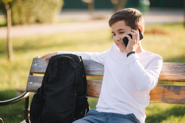 Young boy in white sweatshirt with black backpack sitting on the bench in the park and speak with somebody by the phone