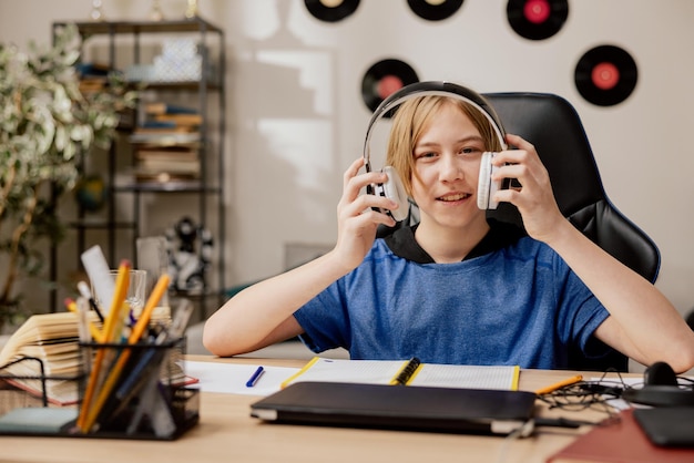 写真 ブロンドの長い髪の少年は、現代のメンズルームのオフィスの机に座っていますティーンエイジャーは宿題をしていますp