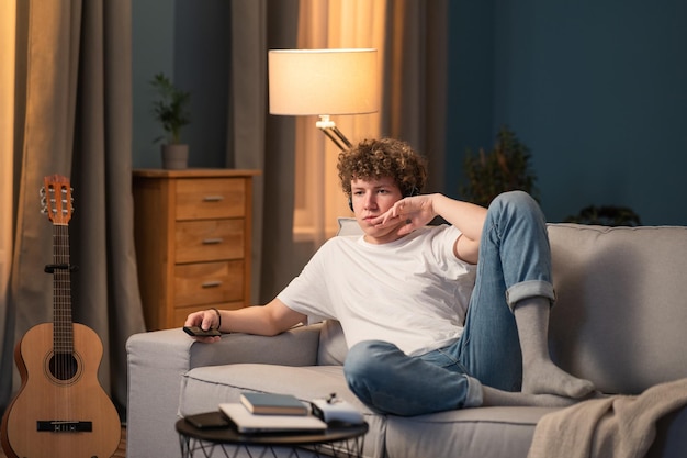 A young boy with curly hair is lying on the couch in homemade clothes with a remote control in hand and watching TV The teenager looks curious about the evening's program