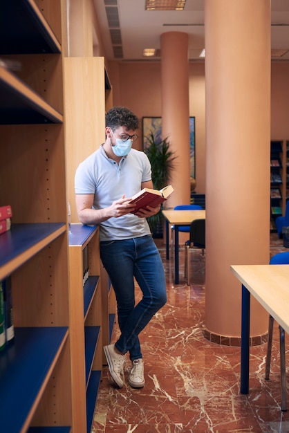 A young boy with a mask is standing checking out a book in the library