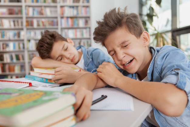 Young boy yawning, falling asleem at the library after studying with his twin brother