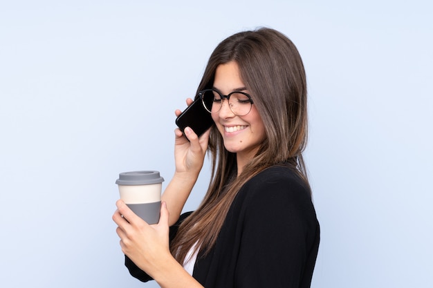 Young Brazilian business woman holding coffee to take away and talking to mobile over isolated blue background