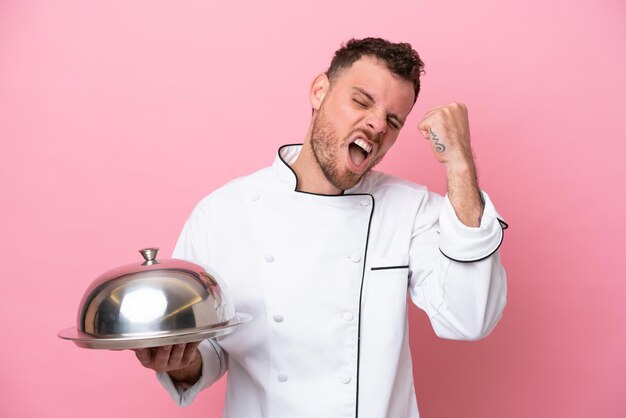 Young Brazilian chef with tray isolated on pink background celebrating a victory