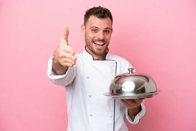 Young Brazilian chef with tray isolated on pink background shaking hands for closing a good deal