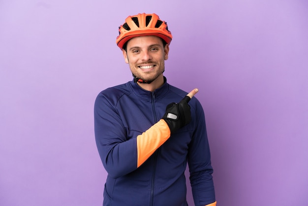 Молодой бразильский велосипедист изолирован на фиолетовом фоне, указывая в сторону, чтобы представить продукт
