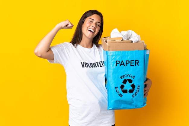 勝利を祝う黄色の背景に分離されたリサイクルする紙でいっぱいのリサイクルバッグを保持している若いブラジルの女の子