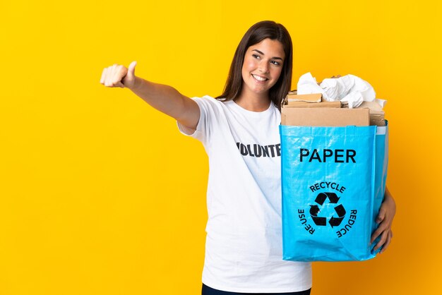 親指を立てるジェスチャーを与える黄色の壁に隔離されたリサイクルする紙でいっぱいのリサイクルバッグを保持している若いブラジルの女の子