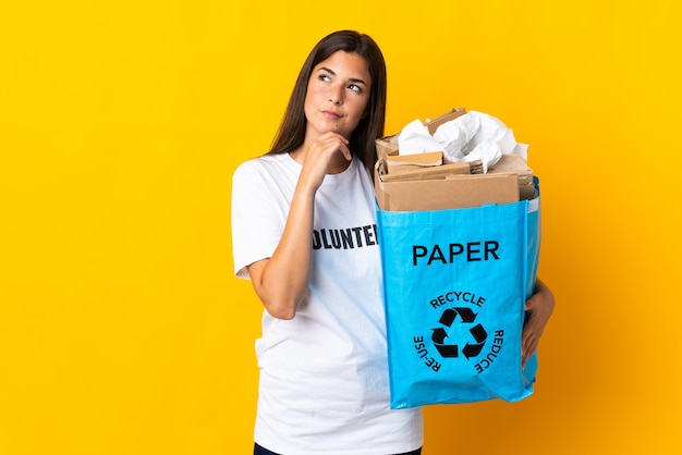 黄色の壁に隔離され、見上げるリサイクルする紙でいっぱいのリサイクルバッグを保持している若いブラジルの女の子