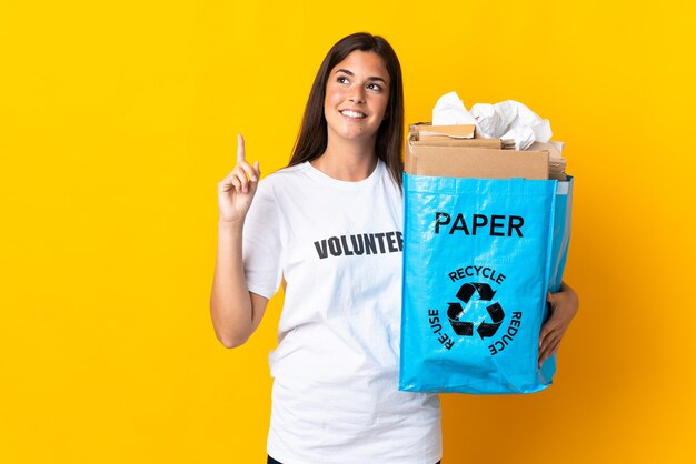 Young brazilian girl holding a recycling bag full of paper to recycle isolated on yellow wall pointing up a great idea