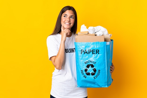 Young brazilian girl holding a recycling bag full of paper to recycle isolated on yellow wall thinking an idea while looking up