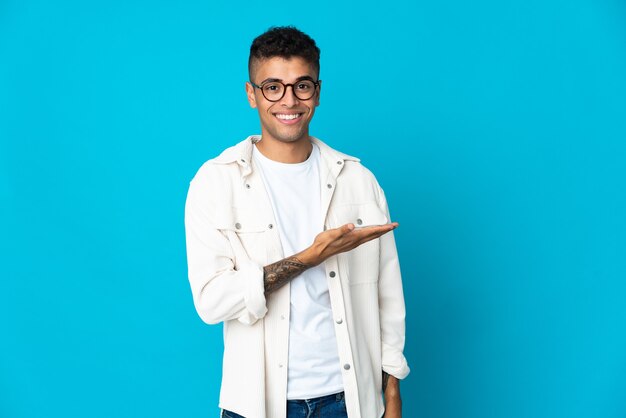 Young Brazilian man on blue background presenting an idea while looking smiling towards