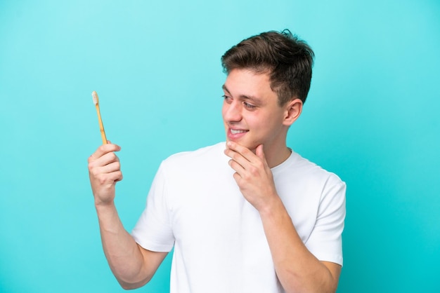 Young Brazilian man brushing teeth isolated on blue background looking to the side and smiling