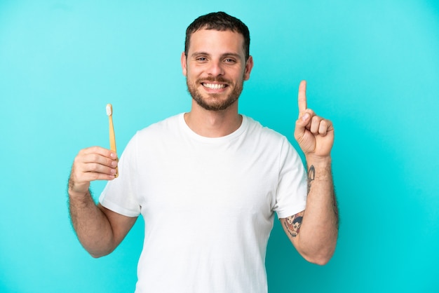 Young Brazilian man brushing teeth isolated on blue background showing and lifting a finger in sign of the best