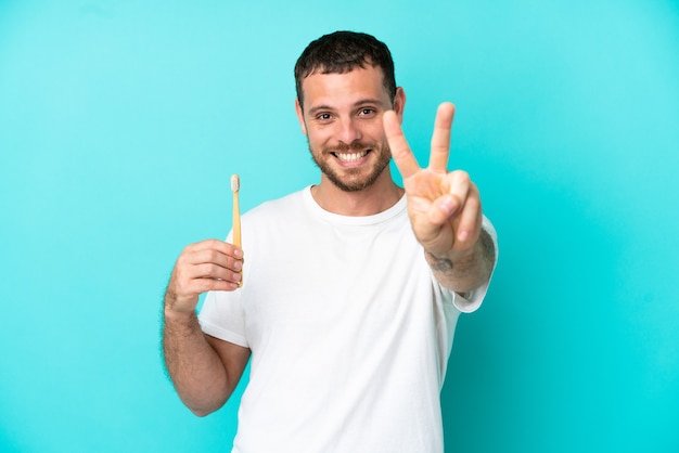 Young Brazilian man brushing teeth isolated on blue background smiling and showing victory sign