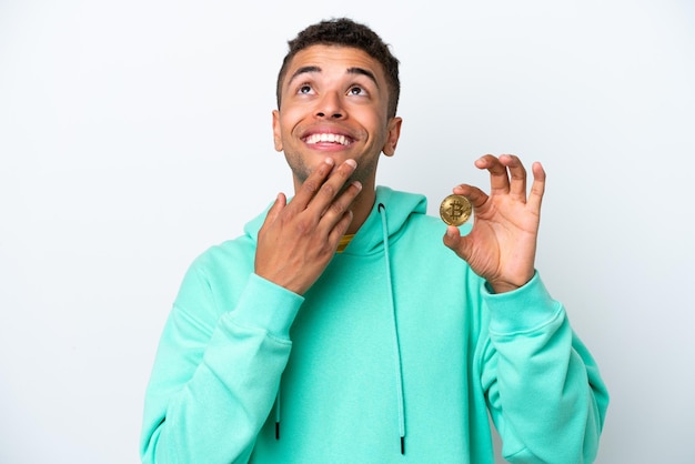 Young Brazilian man holding a Bitcoin isolated on white background looking up while smiling