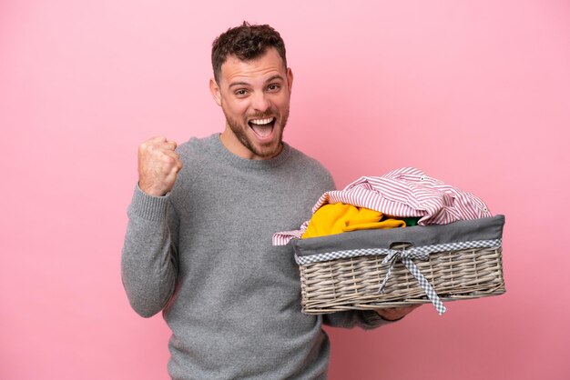 勝者の位置での勝利を祝うピンクの背景に分離された服のバスケットを保持している若いブラジル人男性