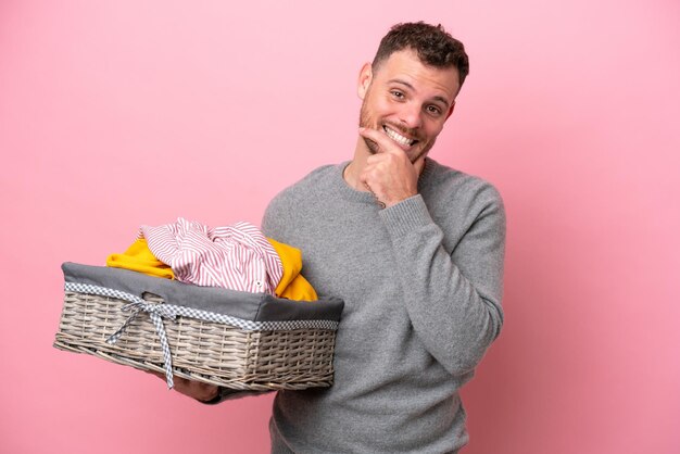 Young brazilian man holding a clothes basket isolated on pink background happy and smiling