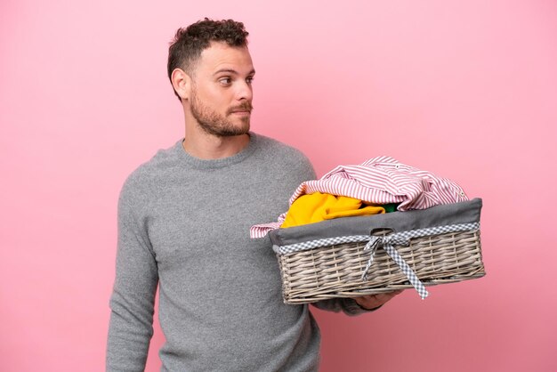 ピンクの背景で隔離の服のバスケットを持って横を見て笑っている若いブラジル人男性