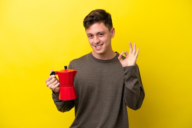 Young Brazilian man holding coffee pot isolated on yellow background showing ok sign with fingers