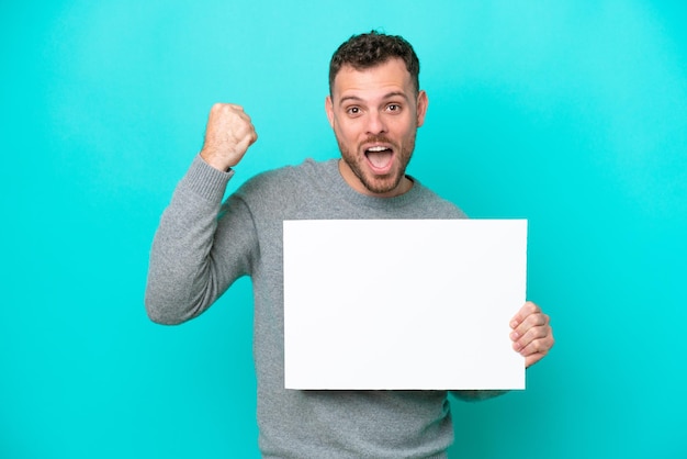 Young Brazilian man holding an empty placard isolated on blue background holding an empty placard and celebrating a victory