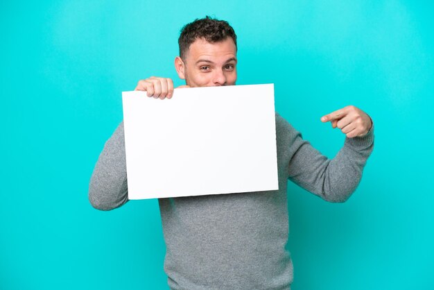 Young Brazilian man holding an empty placard isolated on blue background holding an empty placard and hiding behind it