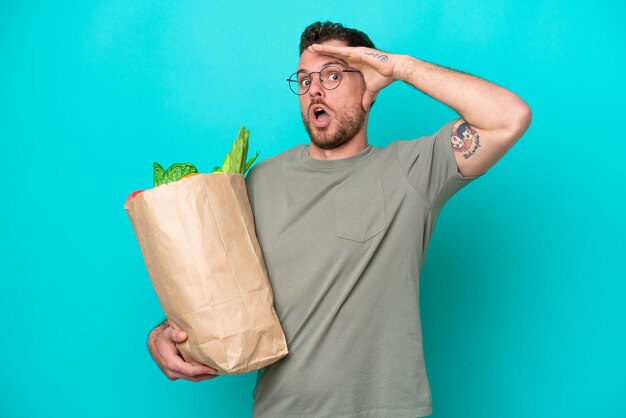 青の背景に食料品の買い物袋を持った若いブラジル人男性が、横を見ながら驚きのジェスチャーをしている