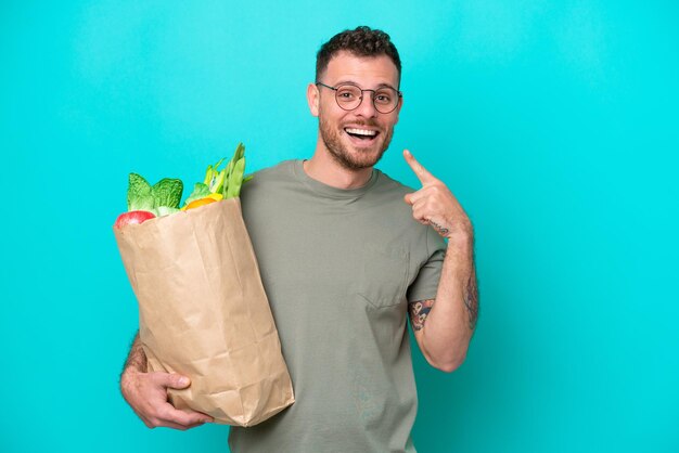 Young Brazilian man holding a grocery shopping bag isolated on blue background giving a thumbs up gesture