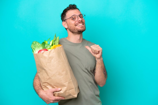 食料品の買い物袋を保持している若いブラジル人男性は、青の背景に誇りを持って自己満足しています