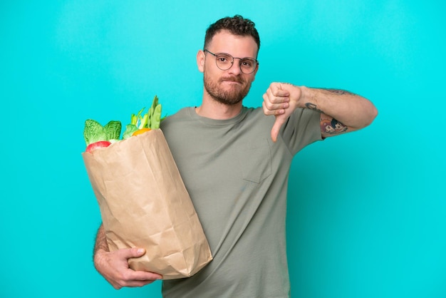 Young Brazilian man holding a grocery shopping bag isolated on blue background showing thumb down with negative expression