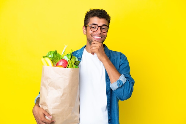 Young brazilian man holding a grocery shopping bag isolated on yellow background looking to the side and smiling