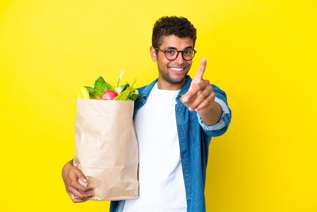 노란색 배경에 격리된 식료품 쇼핑백을 들고 손가락을 들고 있는 젊은 브라질 남자