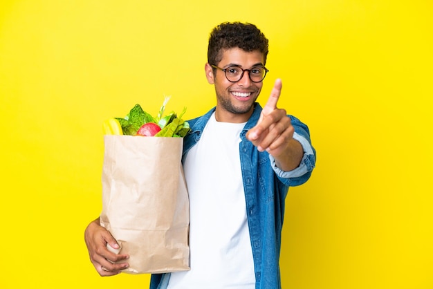 Young Brazilian man holding a grocery shopping bag isolated on yellow background showing and lifting a finger