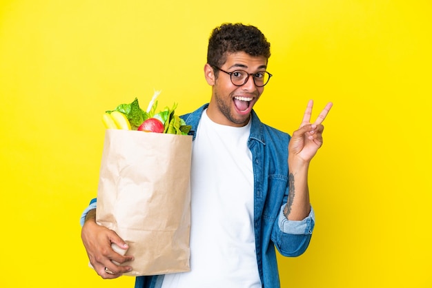 Young Brazilian man holding a grocery shopping bag isolated on yellow background smiling and showing victory sign