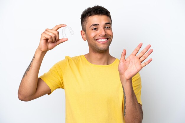 Молодой бразилец с невидимыми брекетами на белом фоне салютует рукой со счастливым выражением лица