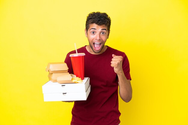 Young brazilian man holding pizzas and burgers isolated background celebrating a victory in winner position