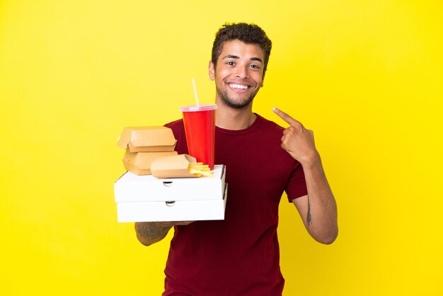 Young brazilian man holding pizzas and burgers isolated background giving a thumbs up gesture