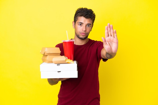 Young brazilian man holding pizzas and burgers isolated background making stop gesture