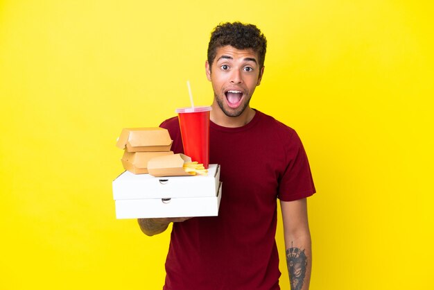 Young brazilian man holding pizzas and burgers isolated background with surprise facial expression