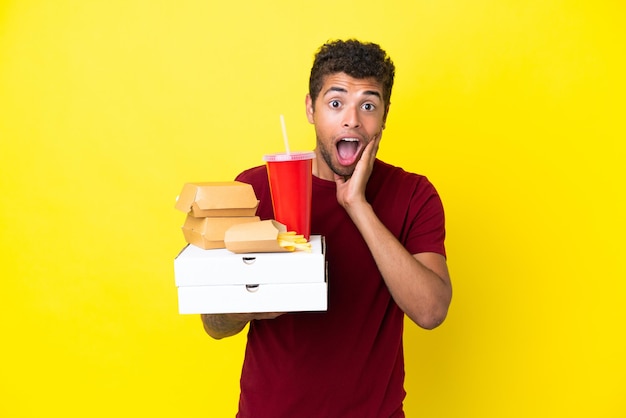 Young brazilian man holding pizzas and burgers isolated background with surprise and shocked facial expression