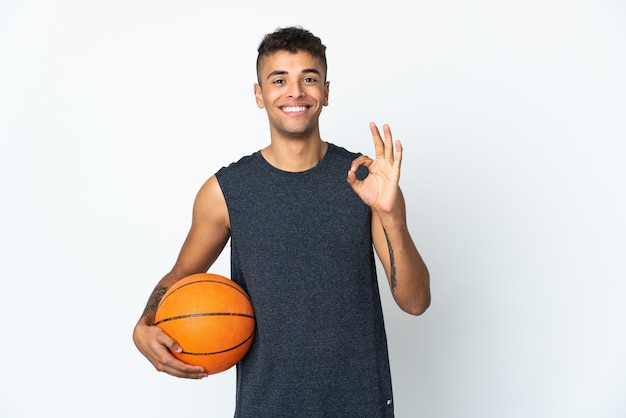 Young Brazilian man over isolated background playing basketball and making OK sign