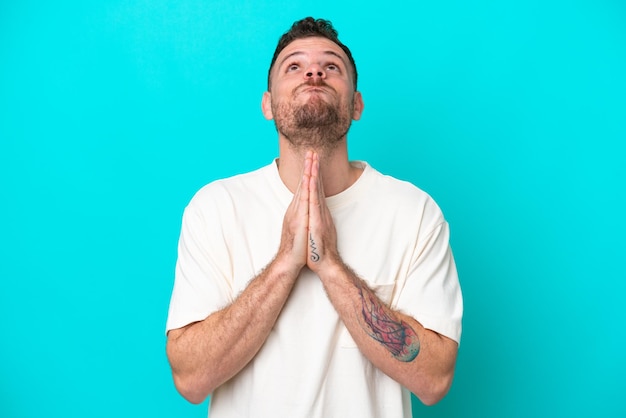 Young Brazilian man isolated on blue background keeps palm together Person asks for something