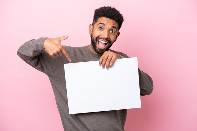 幸せな表情で空のプラカードを保持し、それを指しているピンクの背景に分離された若いブラジル人男性