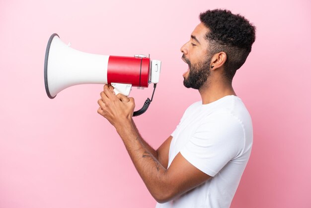 Young Brazilian man isolated on pink background shouting through a megaphone to announce something in lateral position