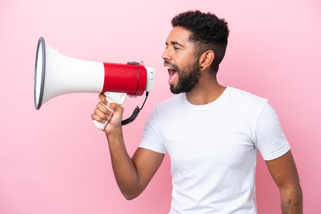 Young Brazilian man isolated on pink background shouting through a megaphone to announce something in lateral position